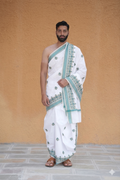 Man wearing a white dhoti with green border against a beige wall.