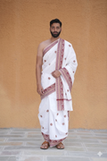 Man wearing a white dhoti with red patterns against a beige wall.