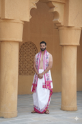 Man wearing a traditional white dhoti with pink border in an architectural setting.